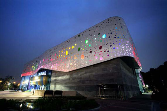 Curved perforated facade illuminated with colored light at night.