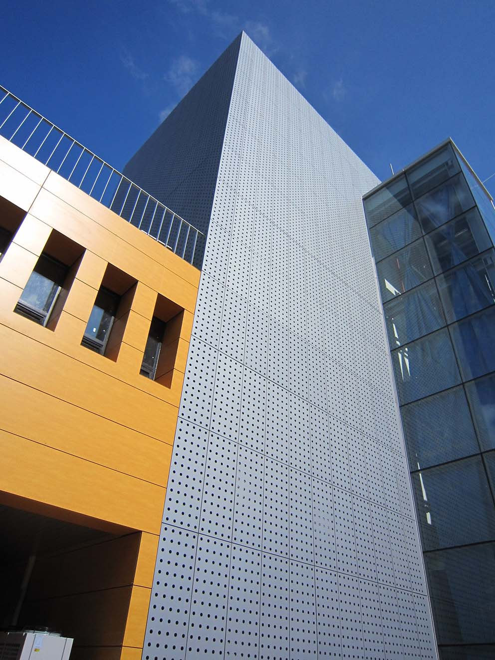 Perforated facade corner meeting warm-colored panels and adjacent glazing, with visible panel joints.