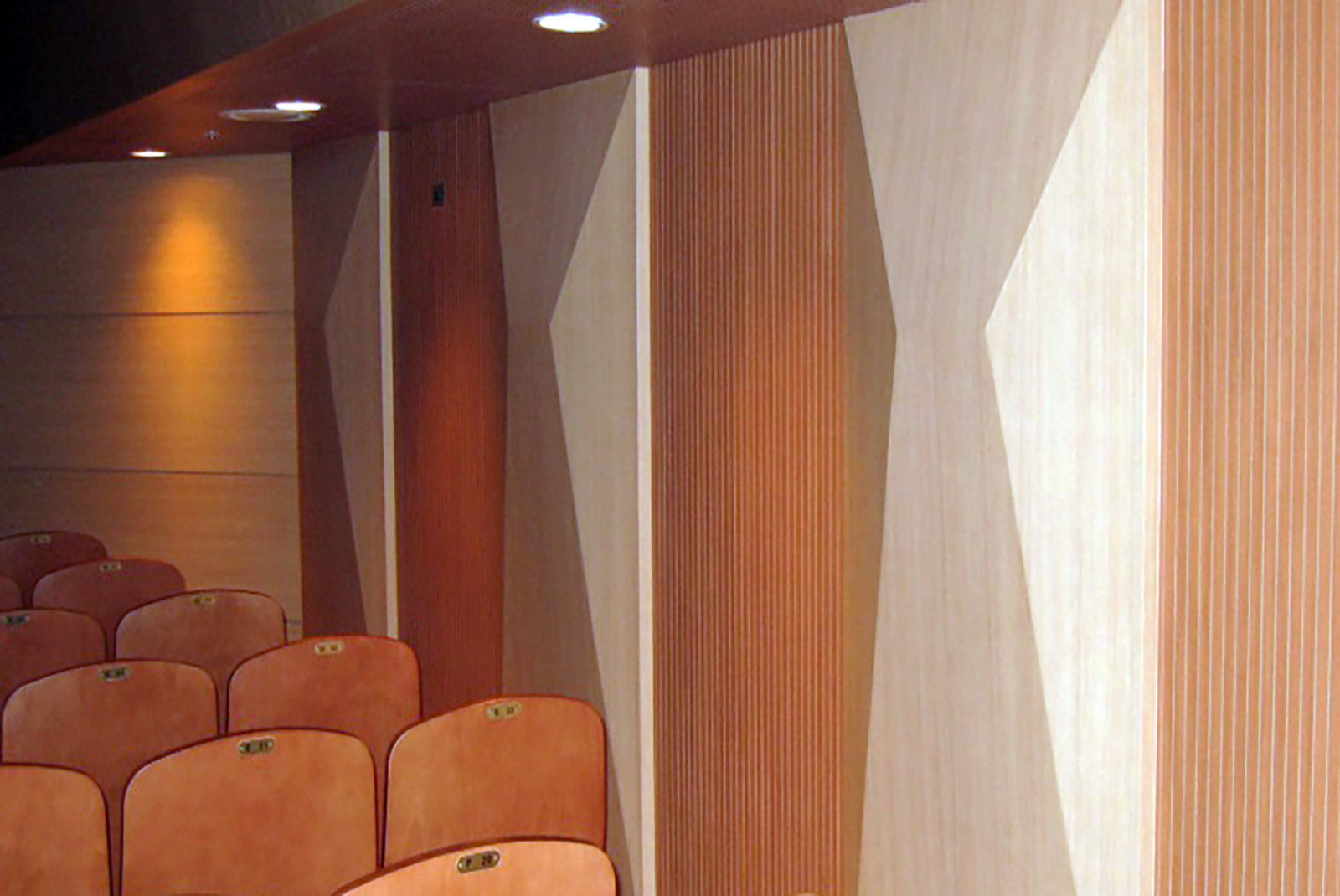 Auditorium seating beside alternating light angled wall panels and warm grooved surfaces beneath ceiling lights.