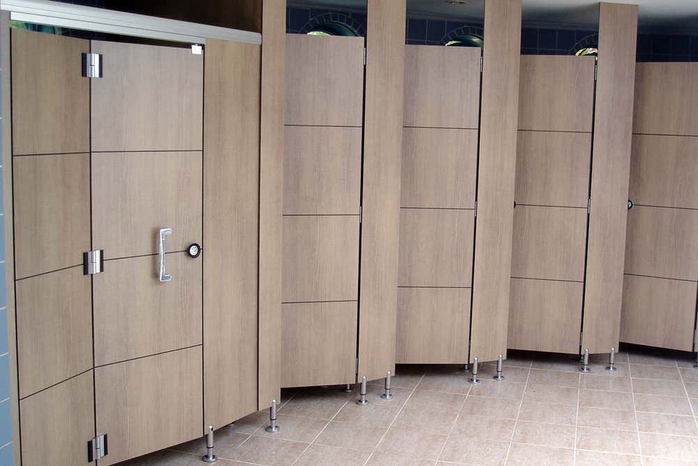 Light wood-tone washroom cubicle doors and partition panels with hinges, handles and floor supports.