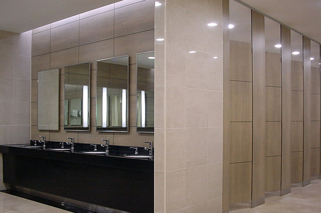 Wood-tone washroom cubicle doors and panels beside a dark vanity and mirrors.