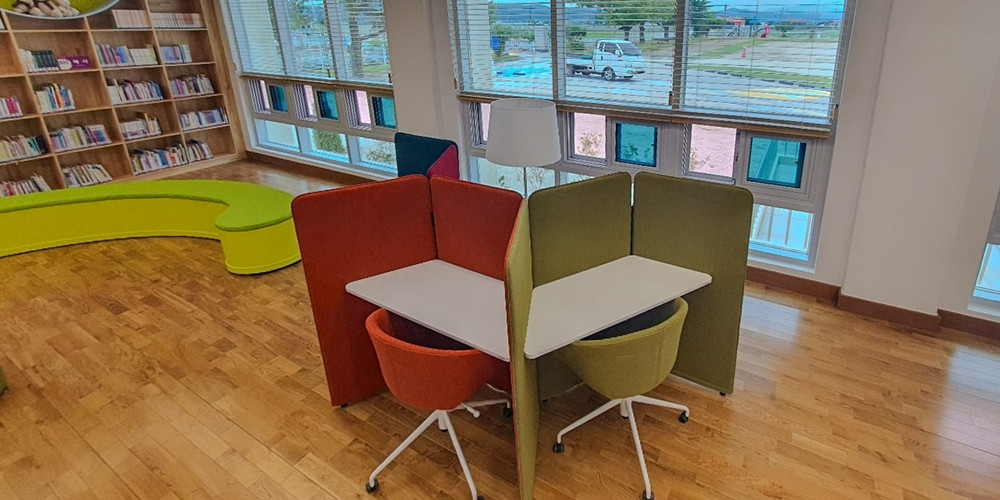 Two pale shaped tabletops within a shared study booth beside school library windows